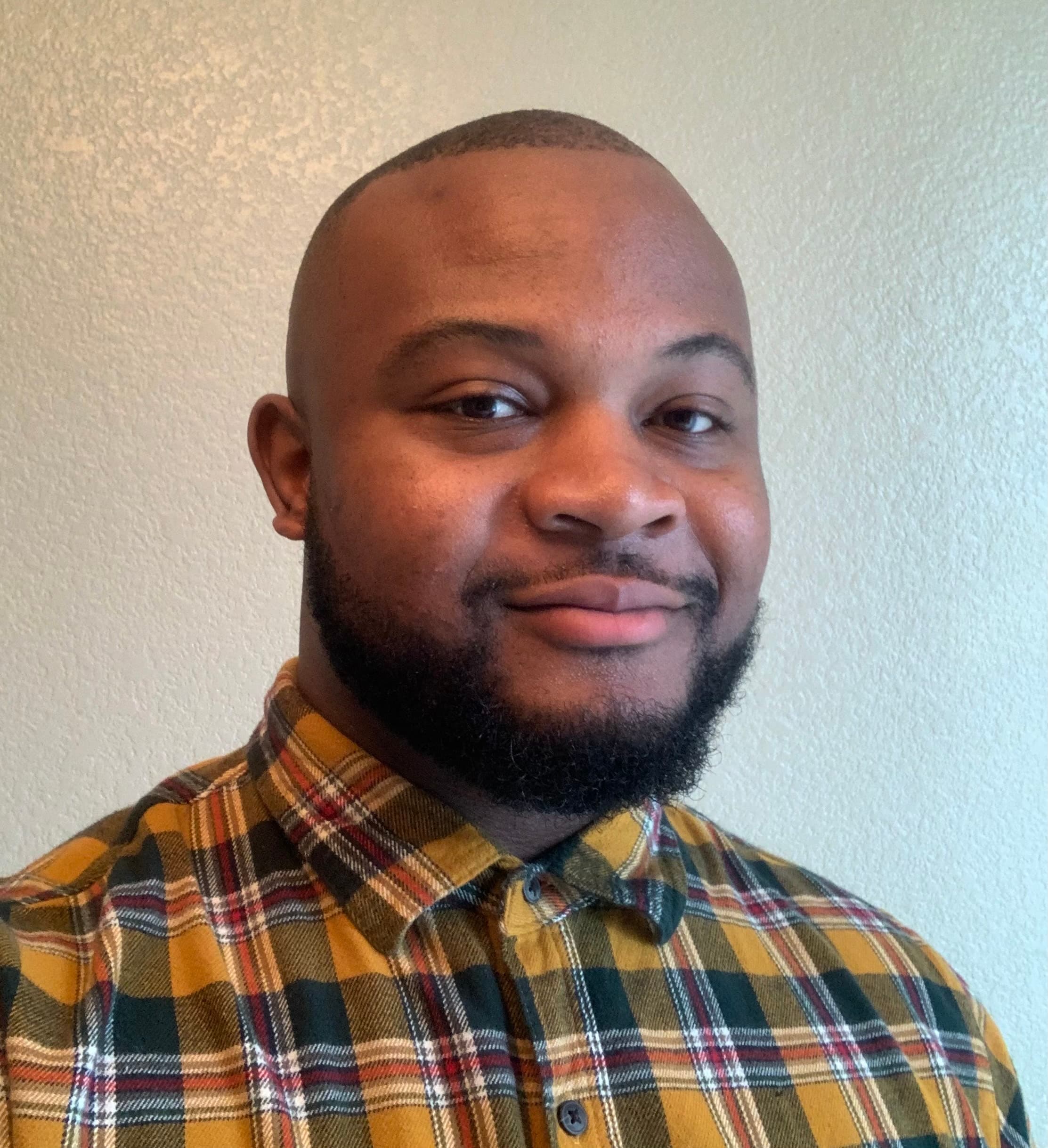 Close-up portrait of a bald man with a beard wearing a plaid flannel shirt, smiling slightly against a plain wall.