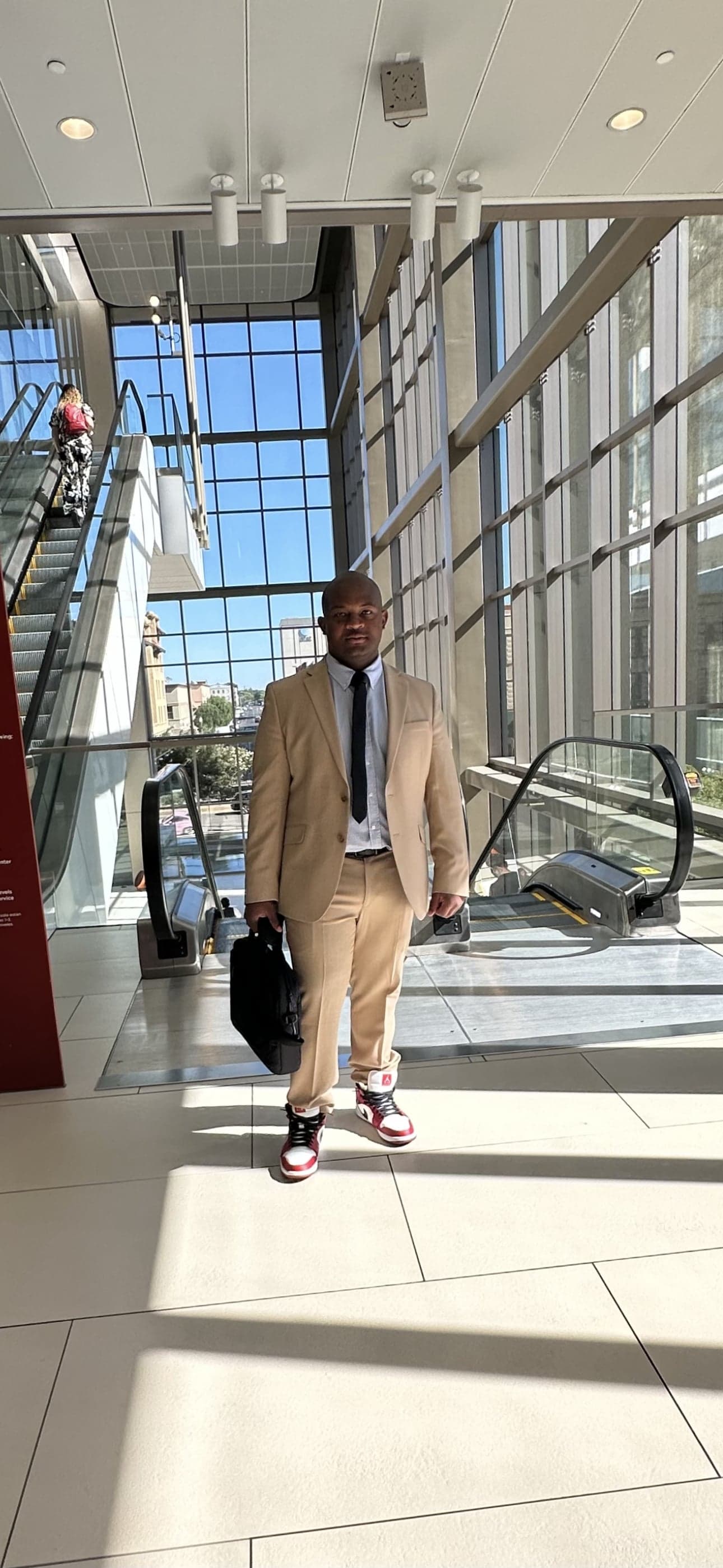 Man in a tan suit carrying a black briefcase, walking through a modern building lobby with escalators and glass walls.
