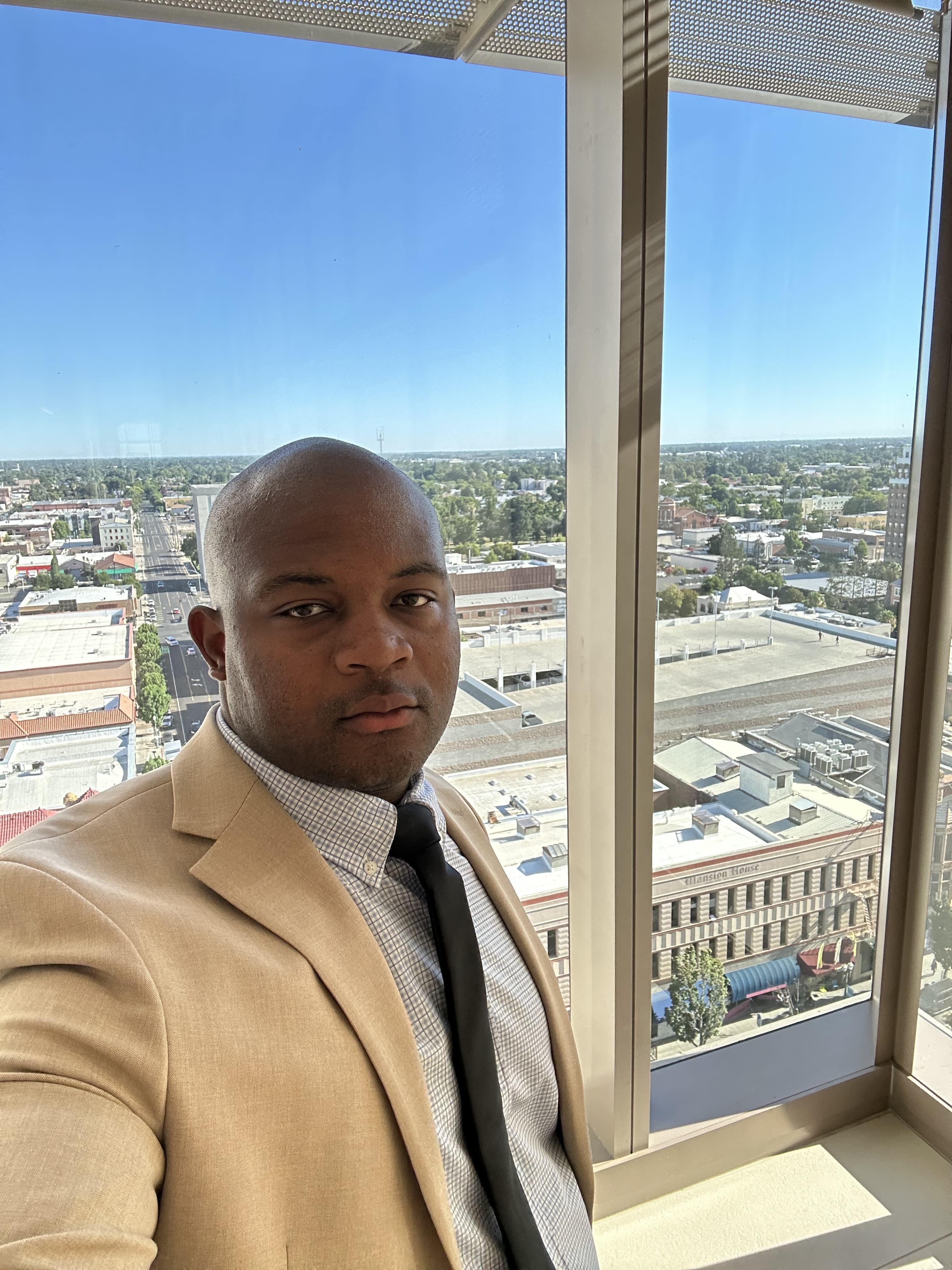 Man in a tan suit posing near a large office window with a city skyline visible outside.