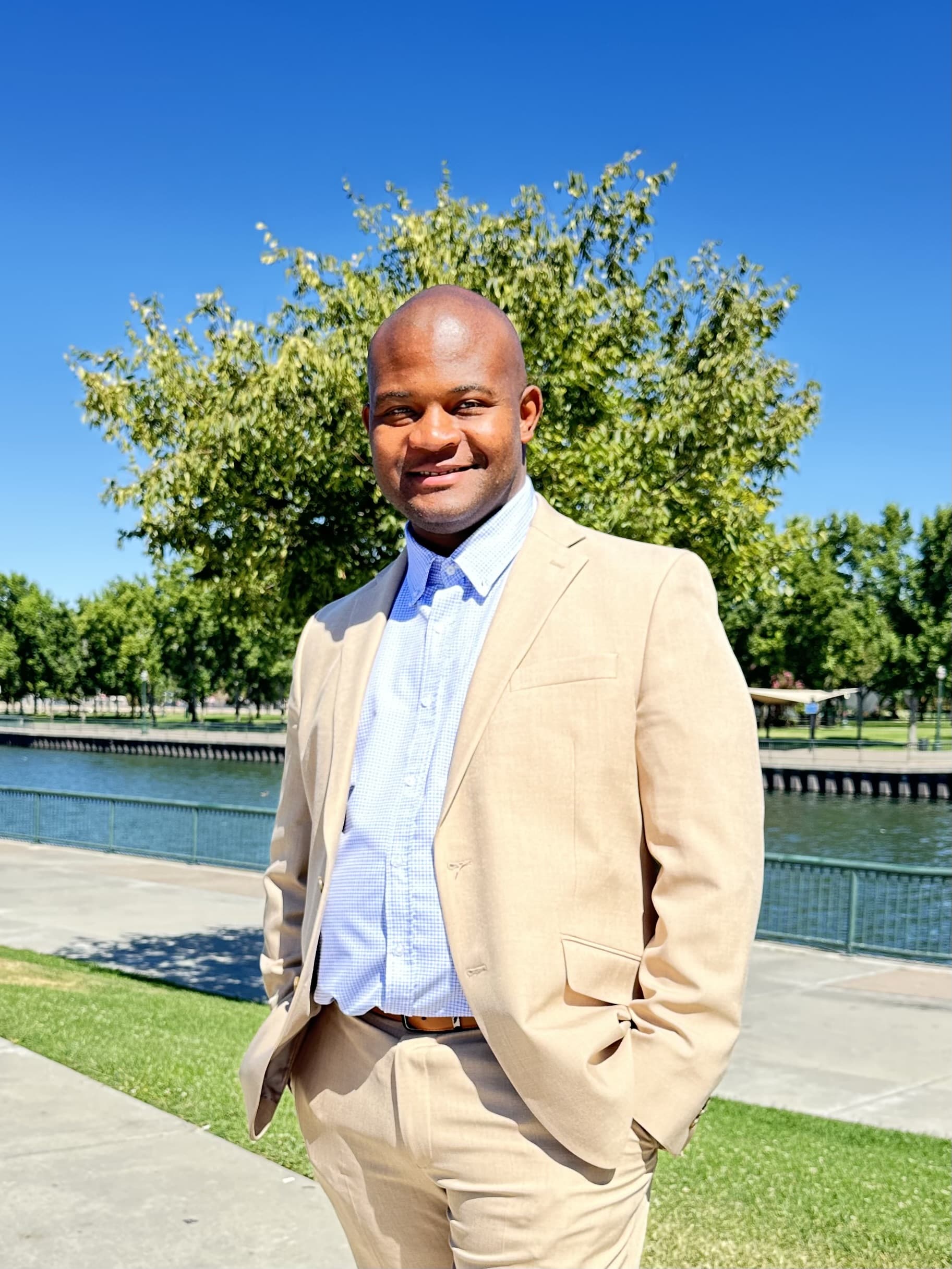 Close-up portrait of a man in a tan suit smiling outdoors with trees and water behind him.
