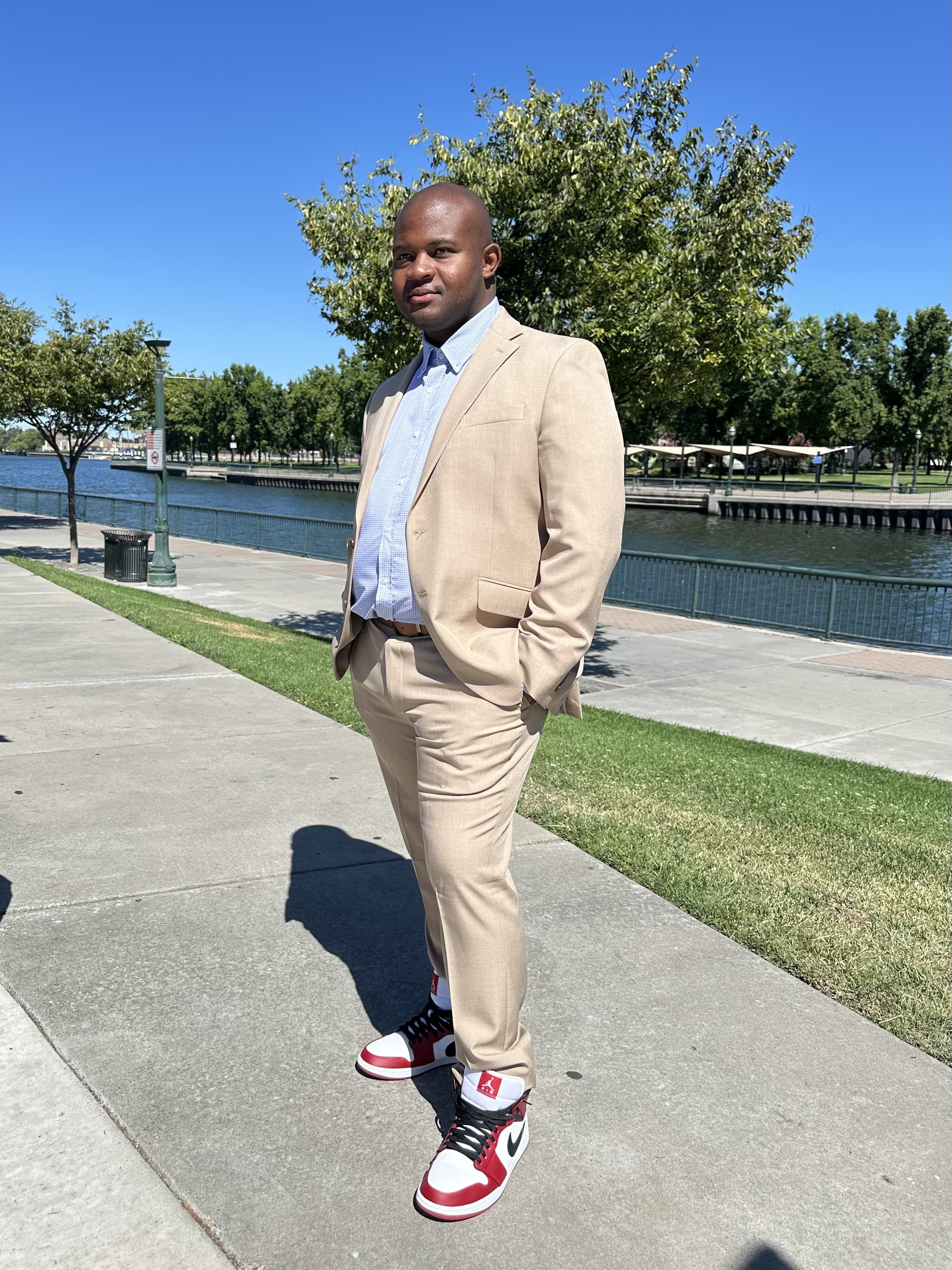 Man in a tan suit with a tie standing on a waterfront promenade, hands in pockets, smiling.