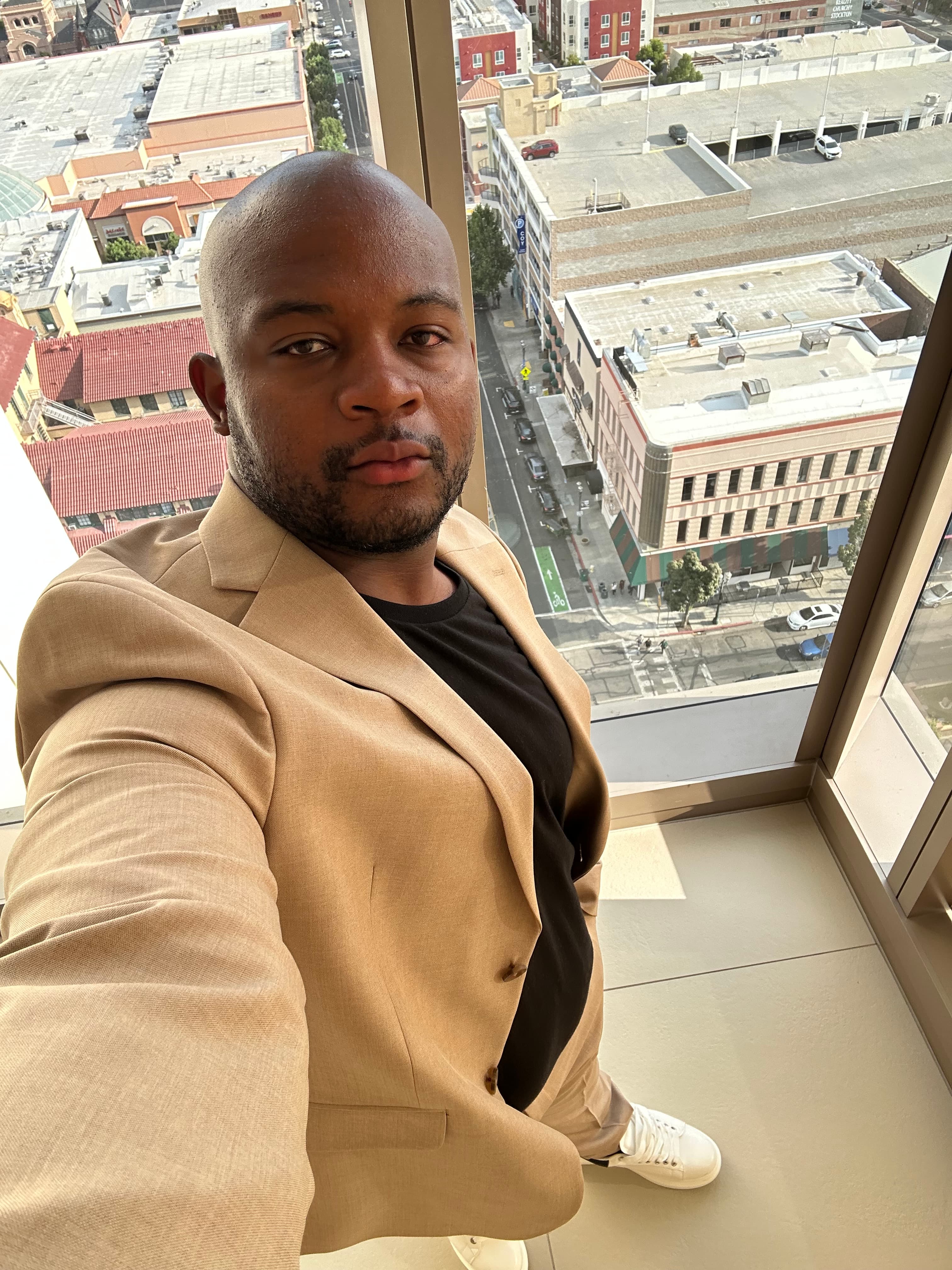 High-angle portrait of a man in a tan blazer, black t-shirt, tan trousers, and white sneakers standing by floor-to-ceiling windows overlooking a city street.