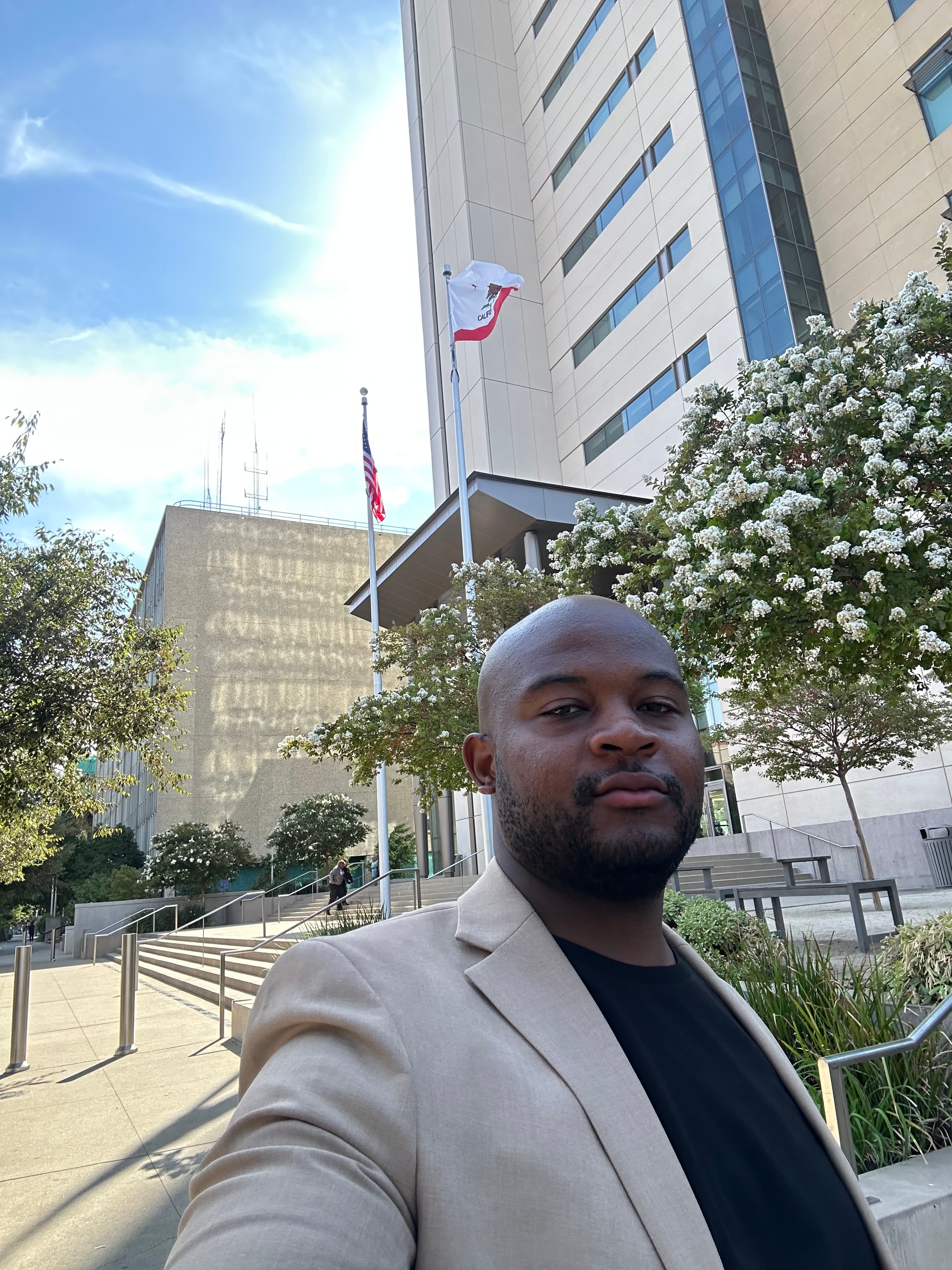 Man in a tan blazer over a black t-shirt standing outdoors in front of modern government buildings, with the California and United States flags on tall poles behind him.