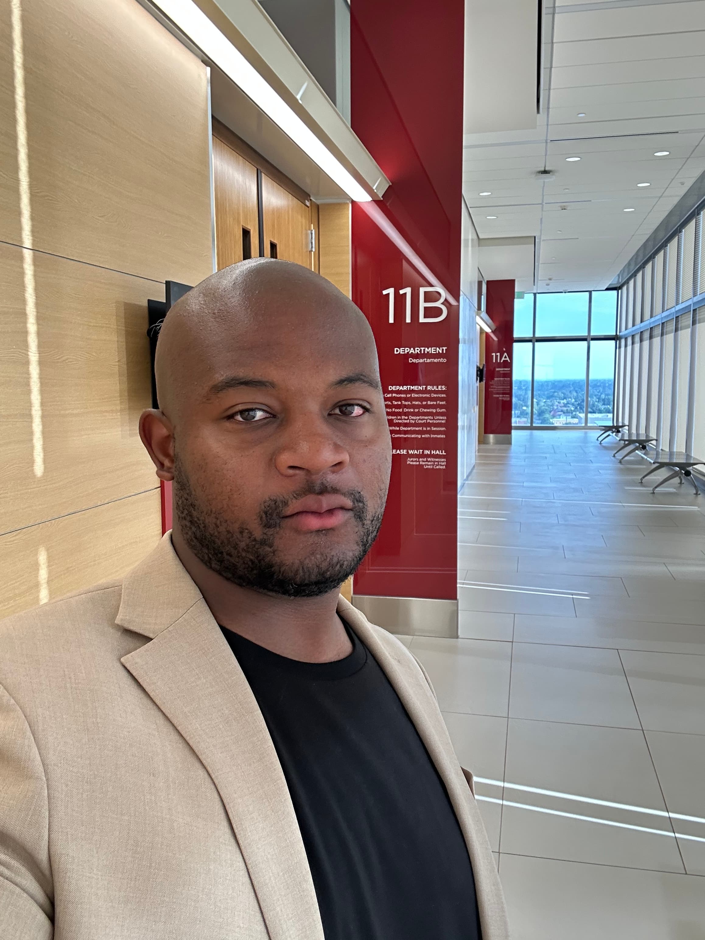 Man in a tan blazer standing in a bright courthouse hallway next to a red pillar labeled 11B Department, with a long tiled corridor and city windows behind him.